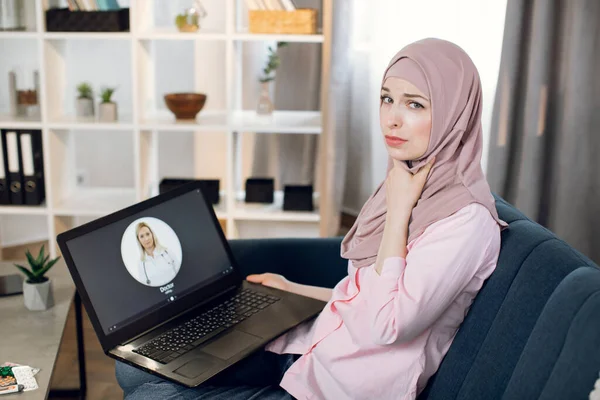 Fáradt kimerült fiatal muszlim nő beteg torokfájással, hív video chat női orvos keresztül egészségügyi alkalmazás, ül a kanapén otthon laptop. Távorvoslás, technológiai koncepció. — Stock Fotó