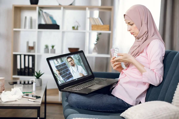 온라인 상의 원격 의사 상담. 히잡을 앓고 있는 젊은 이슬람 여성 이 온라인 상으로 상담을 받고 집에서 노트북을 사용하는 남성 의사와 이야기를 나누며 알약이 묻은 다양 한 물집을 보여 주고 있습니다 — 스톡 사진