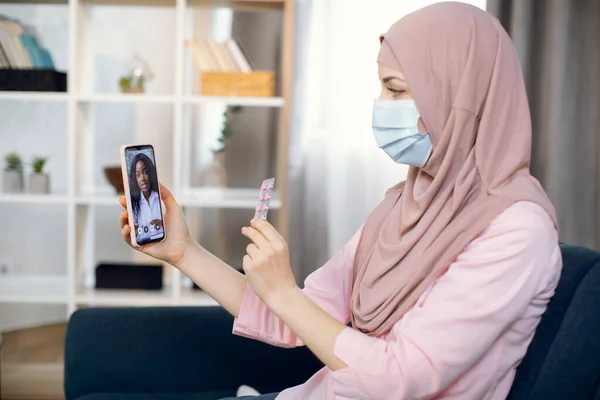 Muzułmańska chora kobieta w hidżabie i masce ochronnej, trzymająca komórkę w jednej ręce i pigułki w drugiej. Afrykańska kobieta lekarz terapeuta na wideokonferencji rozmowy online, pomaga jej pacjentka — Zdjęcie stockowe