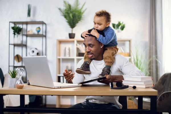 Ocupado afro hombre trabajando en el ordenador portátil con hijo en el cuello — Foto de Stock