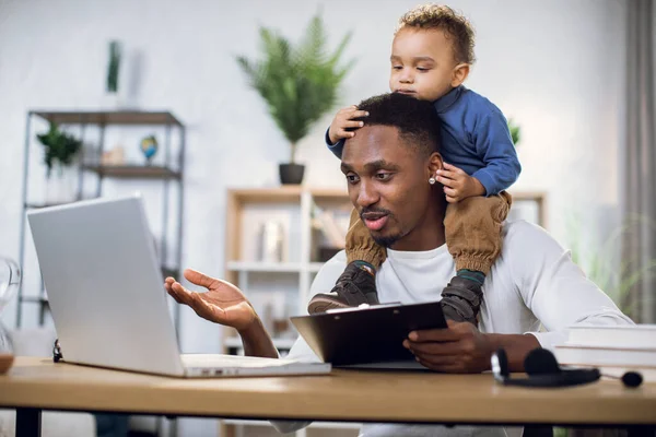 Ocupado afro homem trabalhando no laptop com filho no pescoço — Fotografia de Stock