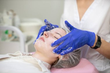 Kadın kozmetik uzmanının eldivenli, profesyonel köpük kullanan kadınların yüzlerini soyarken yakın plan çekimleri, yan görüş. Kozmetoloji kliniğinde akne tedavisi.