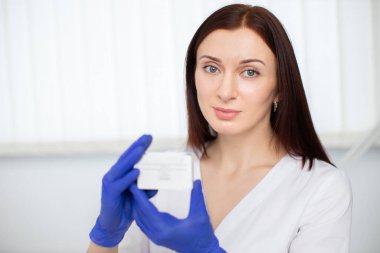 Tıp, kozmetoloji, güzellik ve insan kavramı. Genç profesyonel kadın doktor cildiyecinin portresi, beyaz üniformalı ve mavi eldivenli, ilaç dolu bir kutu tutuyor..