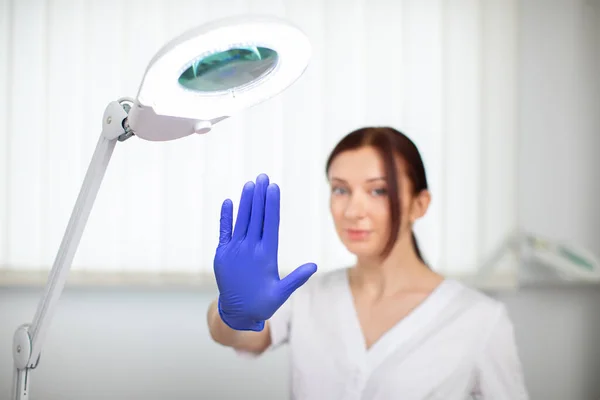 Foto de mulher muito jovem confiante cosmetologist médico em casaco branco e luvas azuis, posando no escritório de beatician luz moderna, mostrando parar gesto. Foco na mão. — Fotografia de Stock