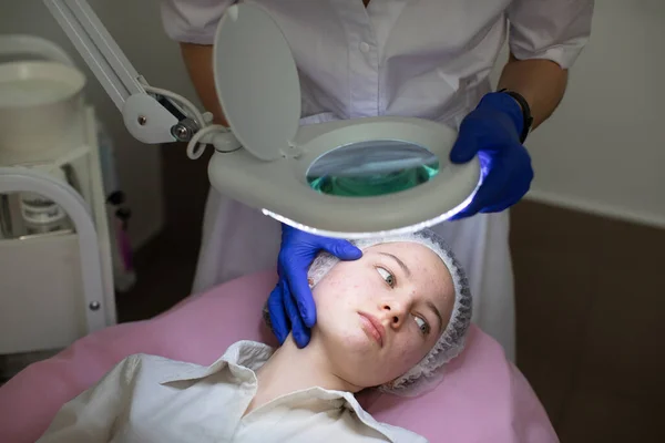 Primer plano de la cara de la joven adolescente con problemas de piel y acné, acostado en la mesa de masaje en la clínica de cosmetología moderna. Manos de doctora esteticista en guantes examina la piel de su paciente. — Foto de Stock