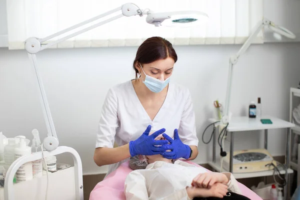 Joven cosmetóloga doctora en máscara protectora y guantes de goma azul hace masaje facial y tratamiento de la piel para clienta joven. Masaje facial spa en salón de belleza, tratamiento de belleza facial. —  Fotos de Stock