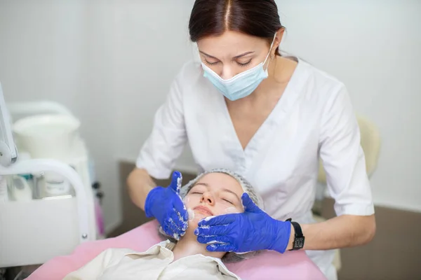 Mujer caucásica joven en máscara facial y guantes haciendo tratamiento facial al cliente en la clínica moderna. Cosmetólogo médico esteticista femenino aplicando polvo limpiador facial en salón de belleza. —  Fotos de Stock