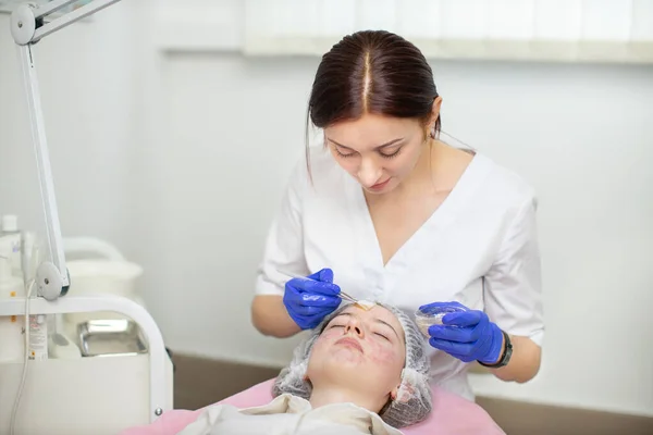 Una dermatóloga cosmetóloga doctora que aplica mascarilla facial en la cara de una joven haciendo cuidado y tratamiento profesional de la piel en el consultorio de cosmetología. Problemas de acné y piel. —  Fotos de Stock