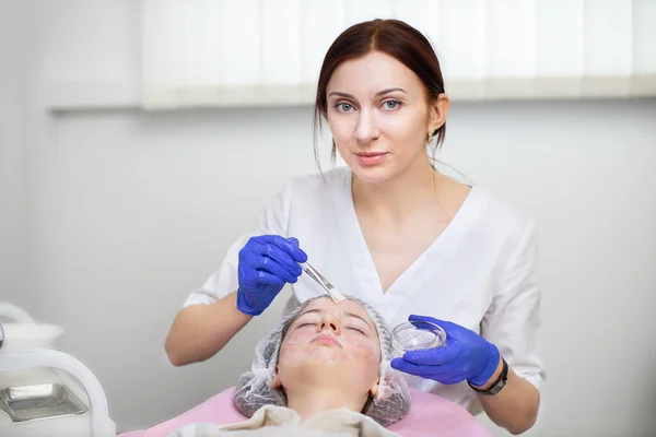 Procedimiento para el cuidado de la piel. Médico femenino en el consultorio de cosmetología aplica mascarilla terapéutica a la cara de la joven adolescente bonita en sombrero desechable, con acné problema de la piel. —  Fotos de Stock