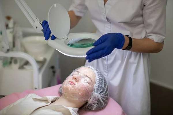 Close-up van jong meisje in professionele kosmetologie kliniek. Gezicht bedekt met plastic folie en masker voor acne behandeling. vrouw schoonheidsspecialiste kijken naar gezicht van klant lloking door lamp. — Stockfoto