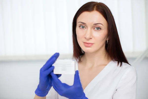 Medicina, cosmetología, belleza y concepto de personas. Retrato de cerca de una joven profesional médico esteticista dermatólogo, en uniforme blanco y guantes azules, sostiene la caja con medicamentos. —  Fotos de Stock
