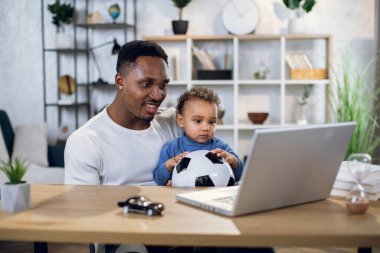 Oğluma bakarken dizüstü bilgisayarda çalışan Afro-Amerikan bir adam.