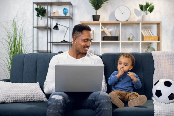 Afro pai sentado com o filho no sofá e trabalhando no laptop — Fotografia de Stock