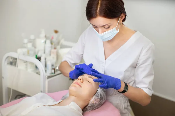 Mujer joven con sombrero médico, acostada en un sofá médico en la oficina de cosmetología. Joven doctora en máscara médica y guantes, limpiándose la frente. Hidratación y rejuvenecimiento de la piel. —  Fotos de Stock