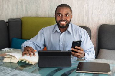 Gülümseyen Afrikalı adam, öğrenci ya da serbest çalışan, sakallı ve gri gömlekli, kafede ya da ortak çalışma alanında oturan, kitap, dijital tablet ve telefondan bilgi arayan.