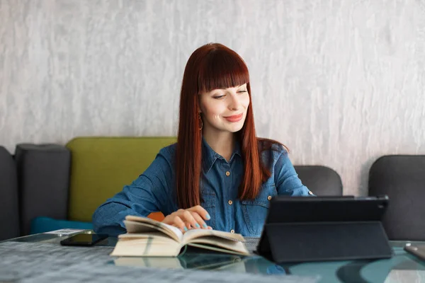 Retrato de joven mujer de pelo rojo caucásico agradable, en camisa vaquera, sentado a la mesa en la cafetería o coworking, leyendo el libro y escribiendo en la tableta pc. Estudio, trabajo freelance, comunicación. —  Fotos de Stock