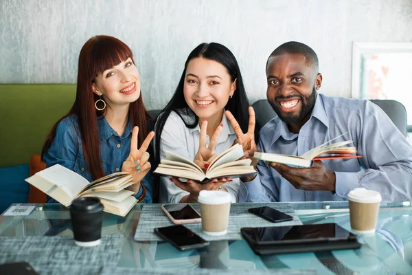 Neşeli üniversite çok ırklı öğrencileri kafede oturup kitaplardan ders çalışmaktan zevk alıyorlar. Genç insanlar kameraya gülümsüyor ve kitap okurken barış zafer işareti gösteriyorlar.. — Stok fotoğraf