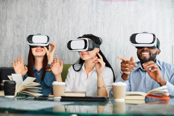 VR experience, futuristic technology. Multiracial three friends moving hands while wearing VR glasses and sitting at cafe. Books and coffee cups on the table