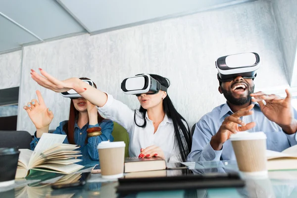 Les jeunes utilisant les nouvelles technologies vr. Multiracial meilleurs amis de collège s'amuser, tout en portant des lunettes de réalité virtuelle dans le café, assis à la table avec des livres et du café. — Photo