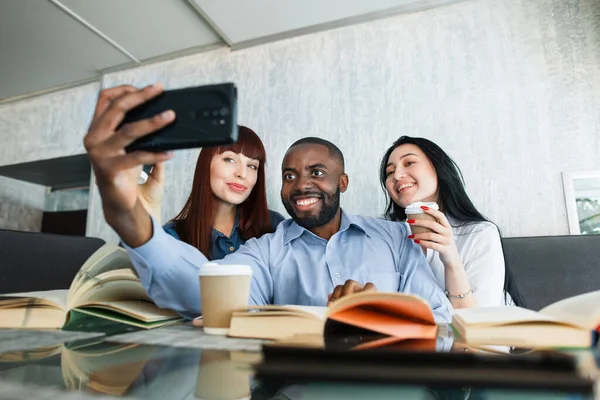 Dersten sonra ya da işten sonra kafede oturup kitaplarla kahve molası veren bir grup mutlu çok ırklı genç arkadaş. Zenci adam, akıllı telefonuyla kız arkadaşlarıyla selfie çekiyor.. — Stok fotoğraf