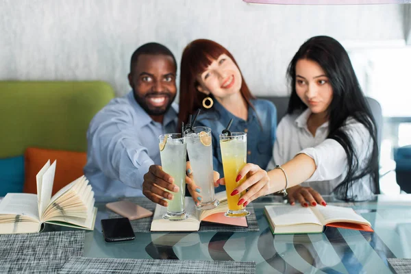 Neşeli üç arkadaş kafede kadeh tokuşturuyor. Afrikalı erkek, beyaz ve Asyalı kadın birlikte ders çalışmaktan, kitap okumaktan ve modern kahve dükkanında meyve suyu içmekten keyif alıyorlar.. — Stok fotoğraf