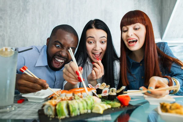 Gente, cibo, divertimento, cena di sushi. Primo piano colpo di eccitati affamati divertenti tre amici multietnici, divertirsi al caffè mangiare cibo asiatico, prendendo rotoli di sushi con bastoncini di bambù tritare. — Foto Stock