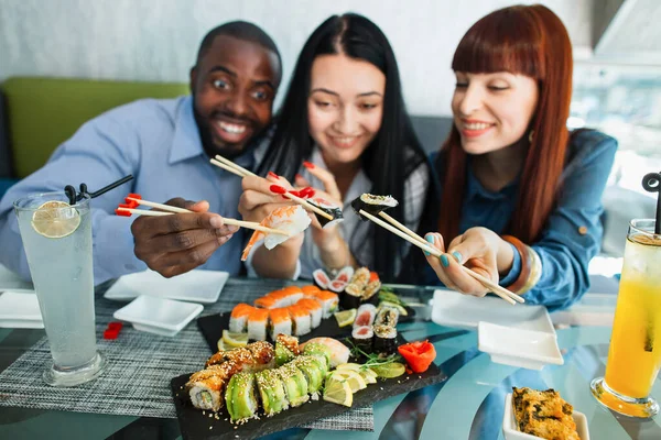 Vista borrosa de la sonrisa agradable y diversa mezcla de amigos de la raza, divirtiéndose y disfrutando del almuerzo en el restaurante chino, sosteniendo palos de madera con deliciosos rollos de sushi. Concéntrate en manos con rollos. — Foto de Stock