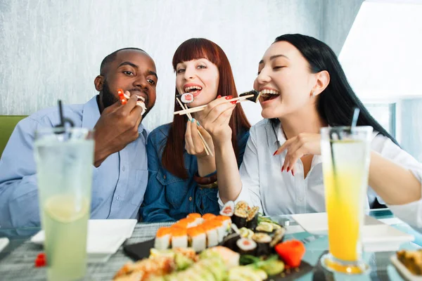 젊고 유쾌 한 다 인종 친구들 이 젓가락으로 초밥을 먹으면서 긍정적 이고 행복해 보이고 아시아인 식당에 앉아 미소짓고 있었다. 사람, 감정, 음식, 우정의 개념. — 스톡 사진