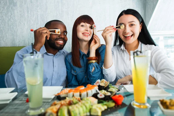 Sushi Time, asian food conception. 세 명의 젊은 다종 민족 친구들즉 아프리카 인 , 코카서스 인 및 아시아 인 소녀들 이 젓가락으로 초밥 조각으로 눈을 가린 모습을 가까이 서 보라. — 스톡 사진