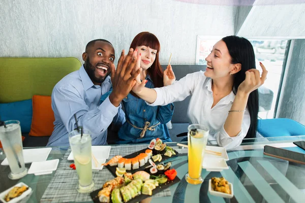 Birlikte harika vakit geçirdik. Mutlu gençler modern kafede suşi rulolarıyla egzotik Asya yemeklerini yiyor ve eğleniyorlar. Asyalı kız ve Afrikalı adam gülüyor ve beşlik çakıyor.. — Stok fotoğraf