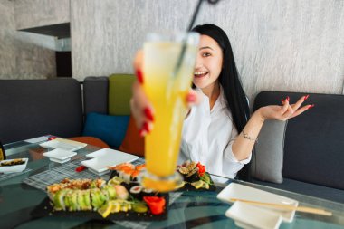 Öğle yemeği ve yaz içkisi zamanı. Taze lezzetli Japon yemekleri. Beyaz gömlekli oldukça heyecanlı bir Asyalı kadın kafede oturuyor, suşi yiyor ve kameraya içki kokteyli gösteriyor. Yüze odaklan.
