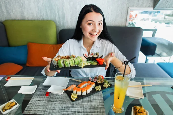 Mladá brunetka asijské žena dívka v ležérní oblečení drží v ruce sushi rolky podávané na černém talíři, sedí v tradiční japonské restauraci. Lidé, životní styl a koncepce asijských potravin. — Stock fotografie