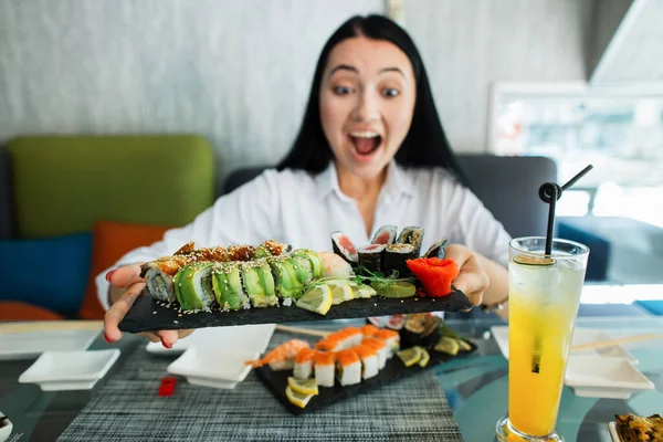 Páni, chutná to. Zblízka vzrušené brunetky asijské ženy šťastný mít vynikající oběd v asijské restauraci a jíst sushi rolky. Zaměřte se na velký černý talíř s barevnými rolky v rukou ženy. — Stock fotografie