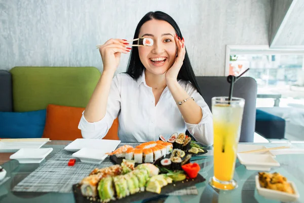Pěkně usměvavá Asiatka sedící u stolu a držící sushi rolku před očima. Krásná mladá žena jí sushi rolku v kavárně. Žena jíst sushi set s hůlkami na restauraci. — Stock fotografie