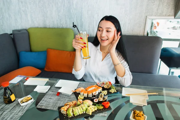 아시아의 젊은 갈색 머리 여자 가 카페에 앉아 유리 테이블에 앉아 신선 한 주스를 마시며 현대사 랑 카페에서 초밥 롤 과 함께 맛있는 점심을 즐긴다. — 스톡 사진
