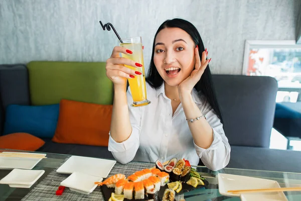 Mladá brunetka hezká Číňanka, sedí u stolu v restauraci, pije exotický ovocný koktejl nebo džus, jí sushi rolky a šťastný úsměv na kameru. — Stock fotografie