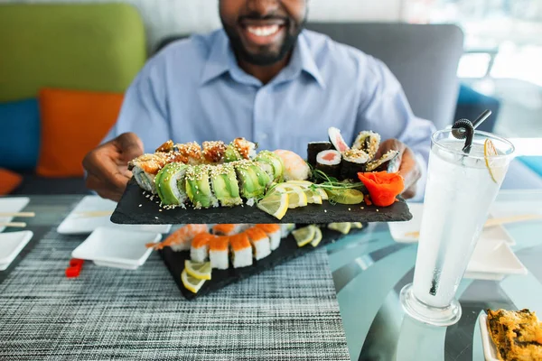 Detailní záběr barevné rozmanité sady sushi rolls na velkém černém talíři v rukou usměvavý mladý Afroameričan muž, sedí u stolu v moderní městské restauraci. — Stock fotografie