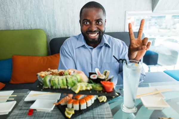 Guapo joven afro hombre, comiendo sushi, sentado a la mesa en el restaurante, sonriendo con la cara feliz, mostrando la paz o la señal número dos. Gente, emociones, comida, concepto de tiempo de sushi. — Foto de Stock
