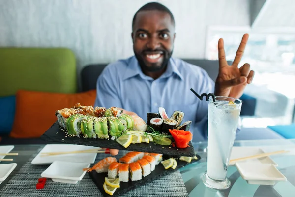 Vista borrosa del guapo chico afroamericano de 30 años, sentado en la cafetería y mostrando a la cámara el signo de paz dos números y un gran plato con un conjunto de deliciosos rollos de sushi. Concéntrate en la placa. — Foto de Stock