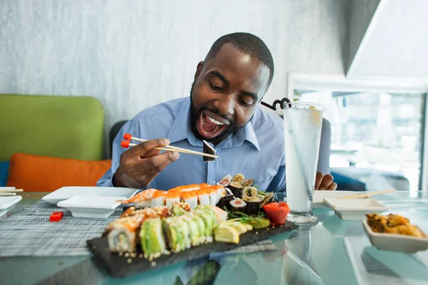Joven hambriento afroamericano emocionado comiendo rollo de sushi caliente usando palillos, sentado a la mesa en la cafetería moderna. — Foto de Stock