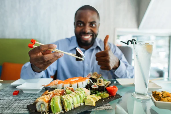 Detailní záběr afrického muže, jak jí sushi v kavárně. Pohledný usměvavý černoch jí sushi rolky, používá hůlky, vede si výborně, palec nahoru prsty. Soustřeď se na jeho ruce. — Stock fotografie