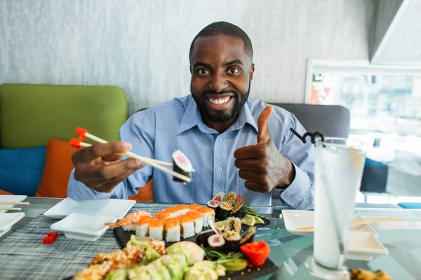 Mladý usmívající se Afroameričan pohledný muž, jedoucí sushi hůlkami, ukazující palec nahoru, dívající se na kameru s veselou a veselou tváří. Muž jíst sushi rolls v orientální kavárně. — Stock fotografie