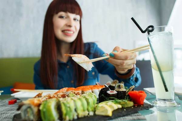 Rozmazaný pohled na sympatickou mladou bělošku, sedící u stolu v restauraci a držící krevety s rýží v dřevěných hůlkách. Sada sushi rohlíků na talíři. — Stock fotografie