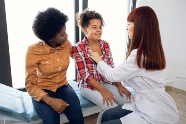 Auscultation, solunum hastalıkları teşhisi ve tedavi. Genç bayan doktor, annesi izlerken Afro-Amerikan kızı muayene ediyor. Hekim alerji uzmanı steteskopla çocuğu inceliyor..