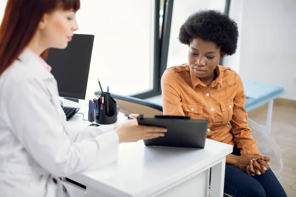 Jeune femme médecin agréable parlant à la patiente afro-américaine, montrant le presse-papiers avec carte médicale et des informations d'assurance. Bilan médical et rendez-vous à la clinique moderne. — Photo