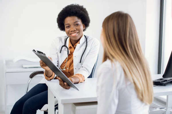 아프리카 계 미국인 여성 의사와 환자와 대화하고 클립보드에 노트를 작성하고 불만 사항을 듣고 개인 정보로 서류 양식을 보여 주고 있습니다. 환자 의등을 통해 보 세요. — 스톡 사진