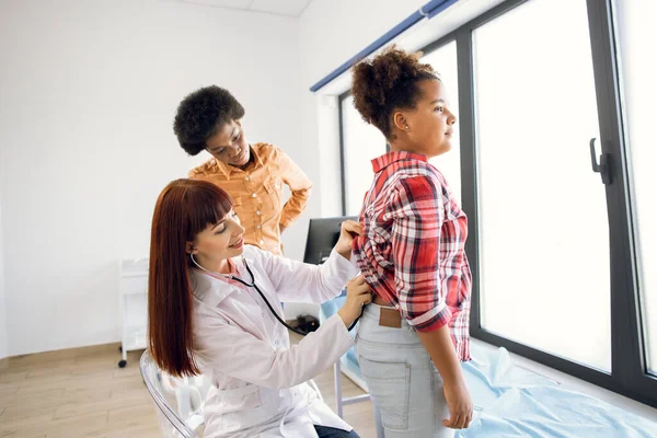 Sintomas de pneumonia e infecção pelo vírus no conceito infantil. Bonito médico gp de 30 anos ouve mestiço menina adolescente respirando com estetoscópio na clínica moderna. Mãe preocupada fica para trás. — Fotografia de Stock