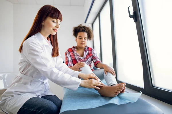 Jovem mulher caucasiana confiante médico ortopedista examinando os pés da estudante afro-americana. Trauma nas pernas e conceito de lesão. Primeiros socorros em pés trauma. — Fotografia de Stock