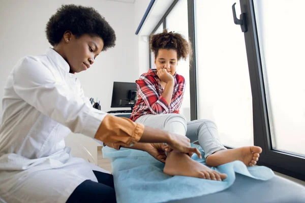 Bonito encaracolado mestiço adolescente de 12 anos menina, sentado no sofá na clínica moderna, enquanto jovem de 30 anos médico africano ortopedista reumatologista examinando pacientes perna. — Fotografia de Stock