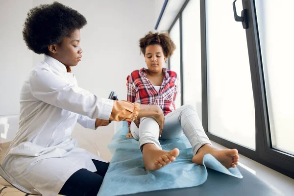 Reabilitação pós-traumática, fisioterapia desportiva, osteopatia. Miúda afro com um joelho dorido no consultório médico. Mulher fisioterapeuta negra examinando o joelho lesionado de sua paciente. — Fotografia de Stock